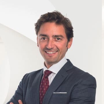 A smiling man in a dark suit and patterned red tie stands against a white background.