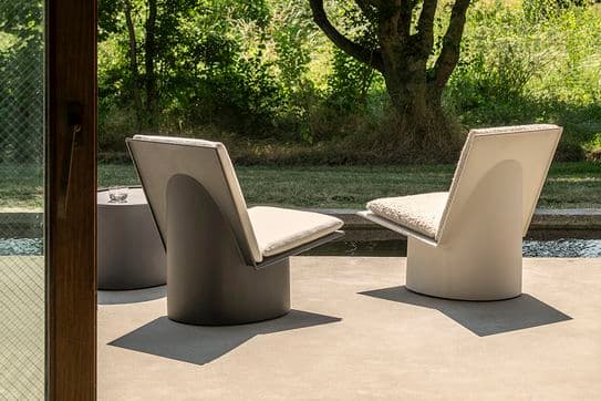Modern patio chairs on a concrete terrace overlooking a lush garden with trees and sunlight casting shadows through a glass door.