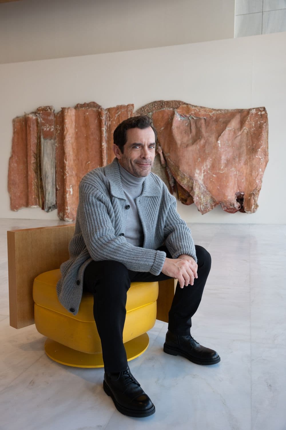 Ένας άνδρας με γκρι ζακέτα κάθεται σε μια κίτρινη καρέκλα μπροστά από μια ανάγλυφη, αφηρημένη τέχνη τοίχου. Φοράει μαύρο παντελόνι και παπούτσια.