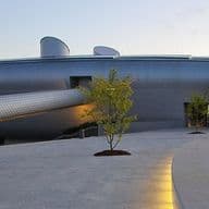 Futuristic building with a metallic exterior, featuring a long, tube-like walkway and surrounded by a landscaped area with young trees.