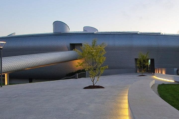 Futuristic building with a sleek, metallic exterior and a cylindrical walkway, surrounded by a landscaped area with small trees.