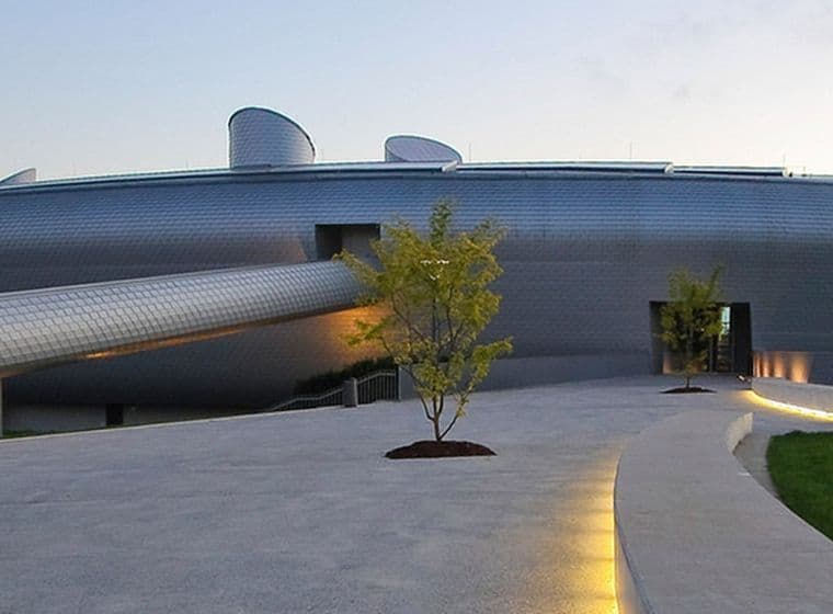 Modern, futuristic building with a cylindrical extension, surrounded by a paved walkway and small trees, under a clear sky.