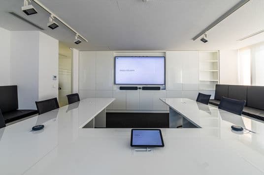 Modern conference room with a large screen, white tables, black chairs, and a tablet on the table. Minimalist design with ample seating.
