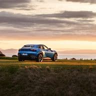 A blue Ferrari Purosangue SUV parked on a grassy hill at sunset, with mountains and water in the background.