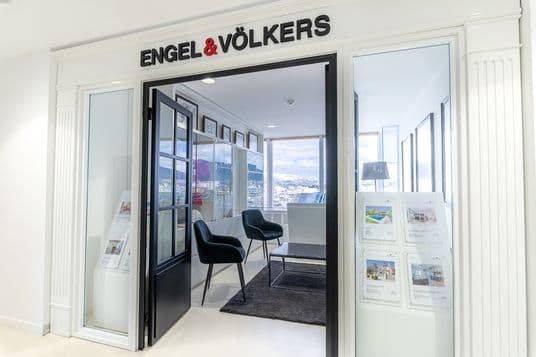 Modern real estate office entrance with Engel & Völkers sign above glass doors, two chairs, a desk, property listings displayed, and large windows showing a city view.