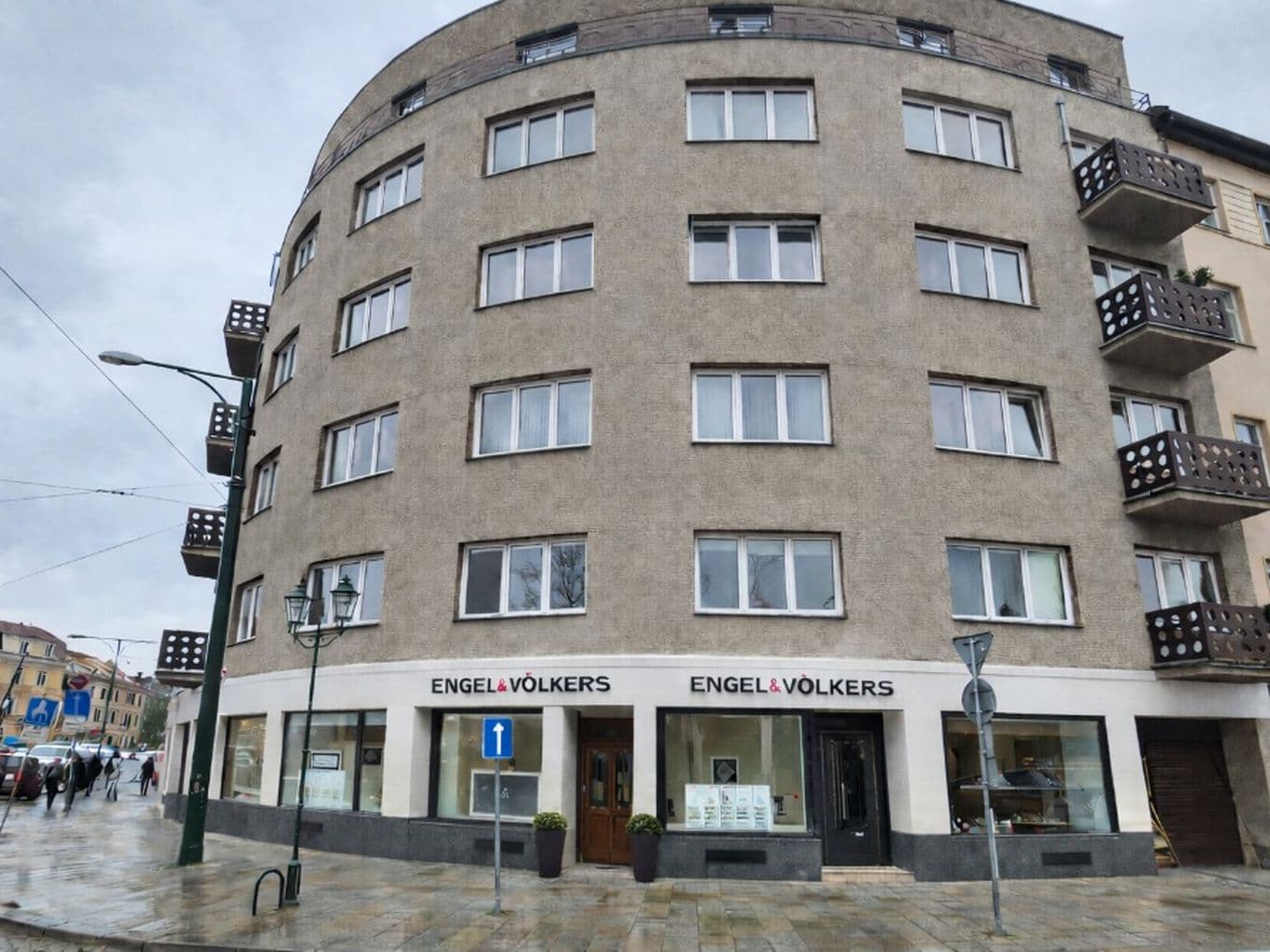 Corner residential building with a rounded façade in the centre of Plzeň. The ground floor features two large glass storefront windows and the entrance to the Engel & Völkers office. A sidewalk and street run in front of the building, with parked cars and street lamps visible.