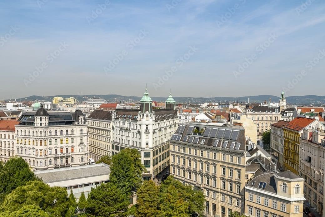 Aerial view of Vienna with historic buildings, red and gray rooftops, and a backdrop of hills under a clear blue sky.