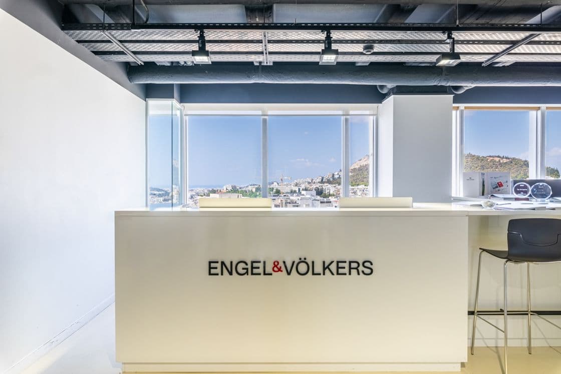Modern office reception with a counter labeled "ENGEL & VÖLKERS," large windows offering a cityscape view, and bar stools on the right.