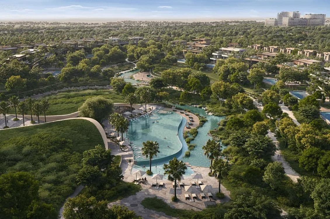 Aerial view of a lush green resort with a large, winding pool, surrounded by trees, pathways, and lounge chairs, under a clear blue sky.