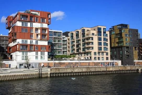 Eine Reihe moderner Wohnungen am Wasser unter blauem Himmel; links ein rot-weißes, mehrstöckiges Gebäude, in der Mitte ein beigefarbenes Glasgebäude, rechts ein dunkles Gebäude mit gelben Akzenten.
