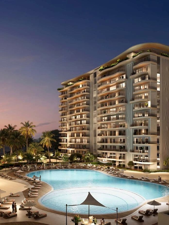 Modern residential towers with curved balconies surround a large central pool at dusk, lined with palm trees and lounge chairs.