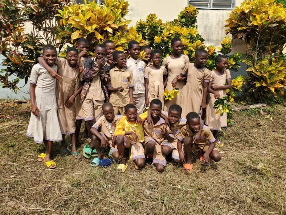 A group of smiling children in school uniforms pose together outside, surrounded by vibrant plants and a beige building.
