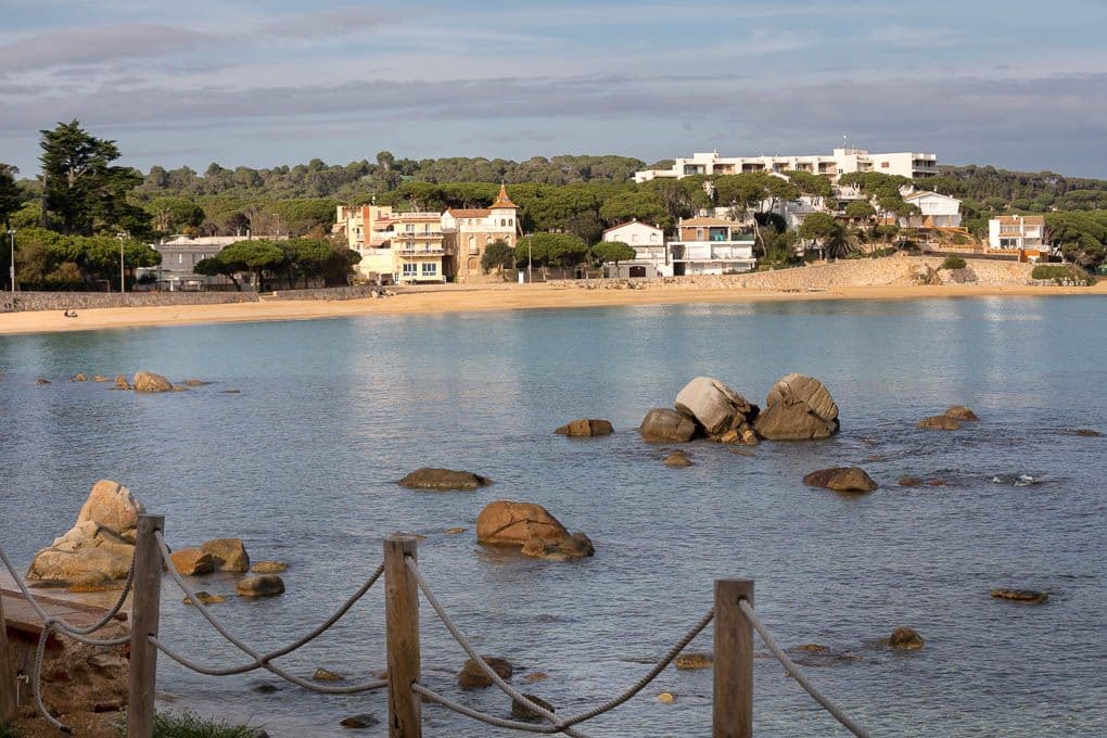 Küstenblick mit Felsen im Wasser, einem Sandstrand und Häusern auf einem Hügel unter einem bewölkten Himmel. Im Vordergrund befindet sich ein Seilzaun.