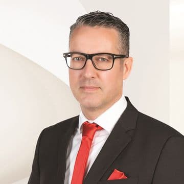 Man with glasses in a dark suit and red tie, standing against a light abstract background.
