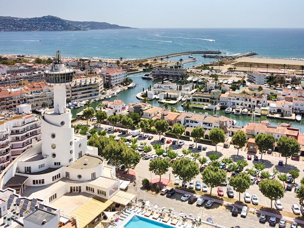 Im Hintergrund sieht man das Meer, dazu Kanäle mit festgemachten Booten und ein Hotel mit Swimmingpool.