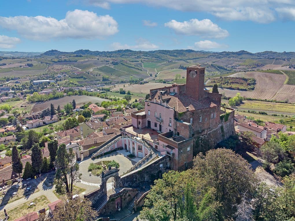 Luftaufnahme einer historischen Burg auf einem Hügel, umgeben von einem Dorf, mit sanften Hügeln und Feldern im Hintergrund unter einem teilweise bewölkten Himmel.