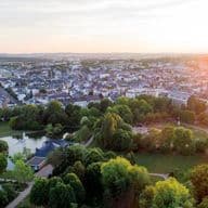 Aerial view of a city with a park in the foreground, featuring a pond with a fountain and a formal garden, all bathed in the warm light of the setting sun.