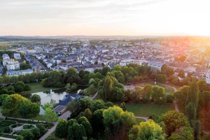 Aerial view of a city with a park in the foreground, featuring a pond with a fountain and a formal garden, all bathed in the warm light of the setting sun.