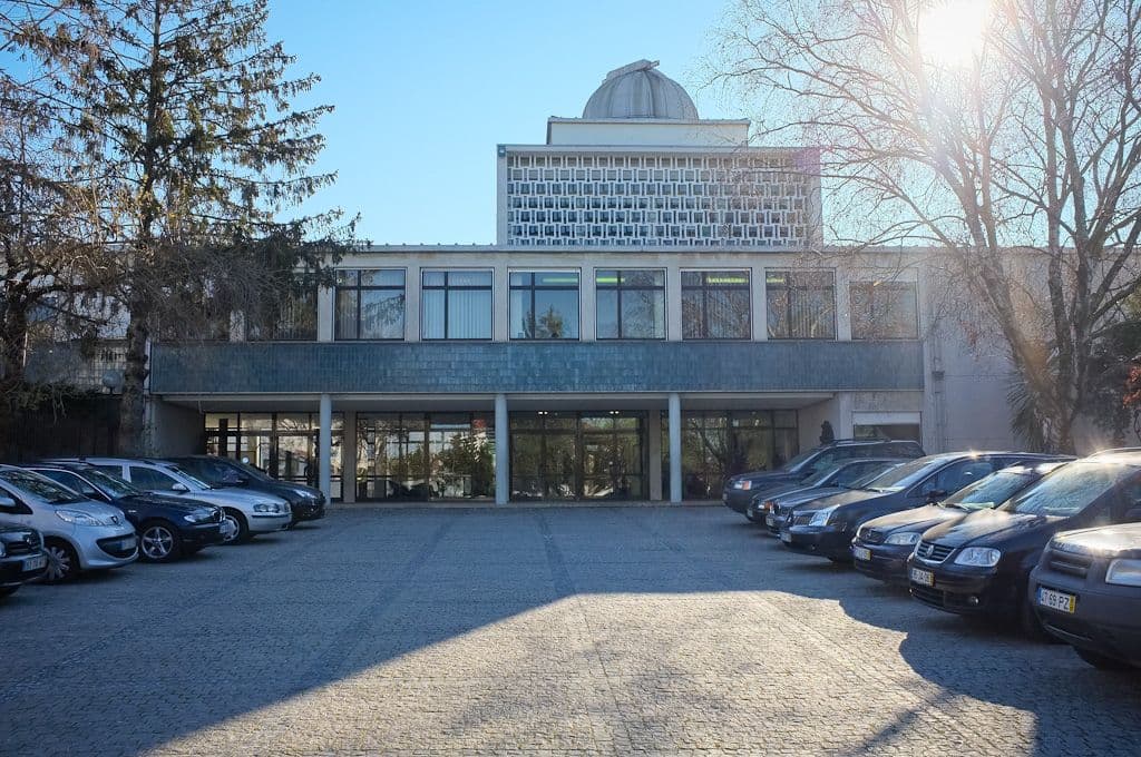 Ein Gebäude mit einer Kuppel, großen Fenstern und einem mit Autos gefüllten Parkplatz an einem sonnigen Tag. Bäume rahmen die Szene ein.