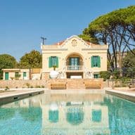 Elegant neoclassical villa with a private swimming pool, surrounded by lush greenery and tall pine trees under a clear blue sky.