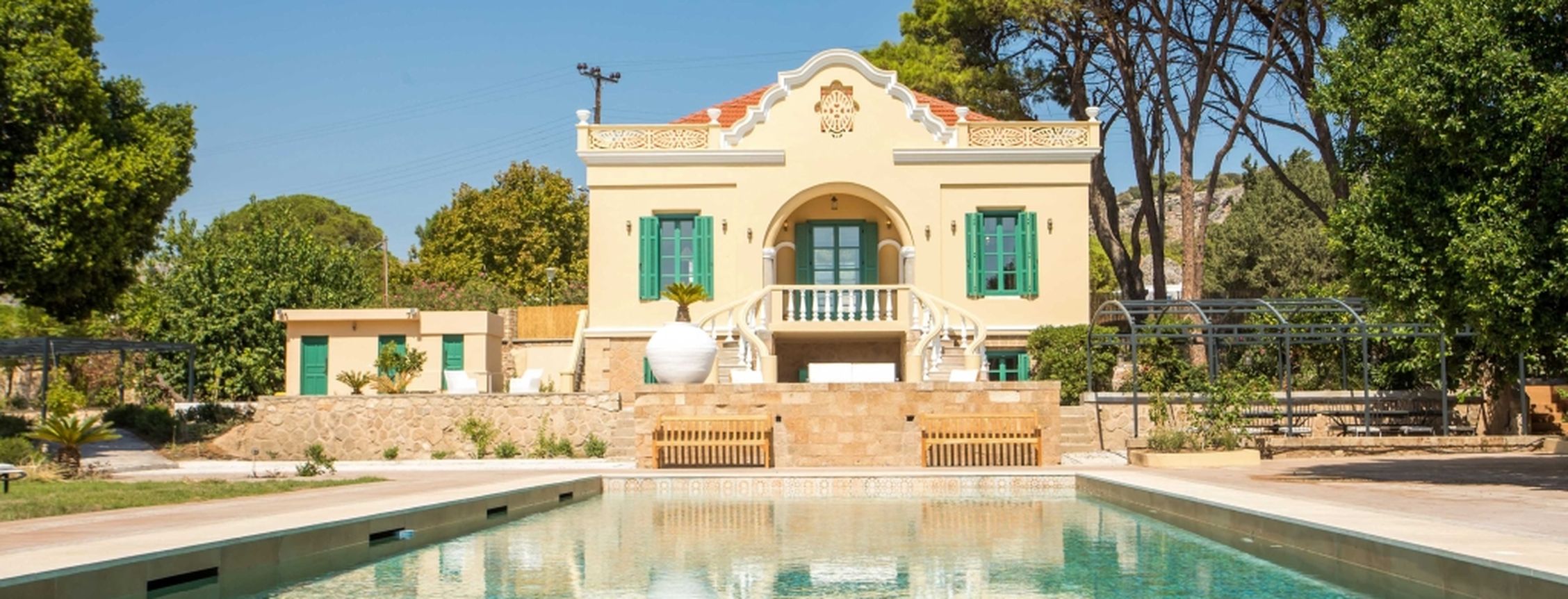 Elegant neoclassical villa with a private swimming pool, surrounded by lush greenery and tall pine trees under a clear blue sky.