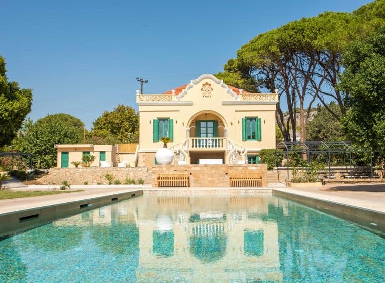 Elegant neoclassical villa with a private swimming pool, surrounded by lush greenery and tall pine trees under a clear blue sky.