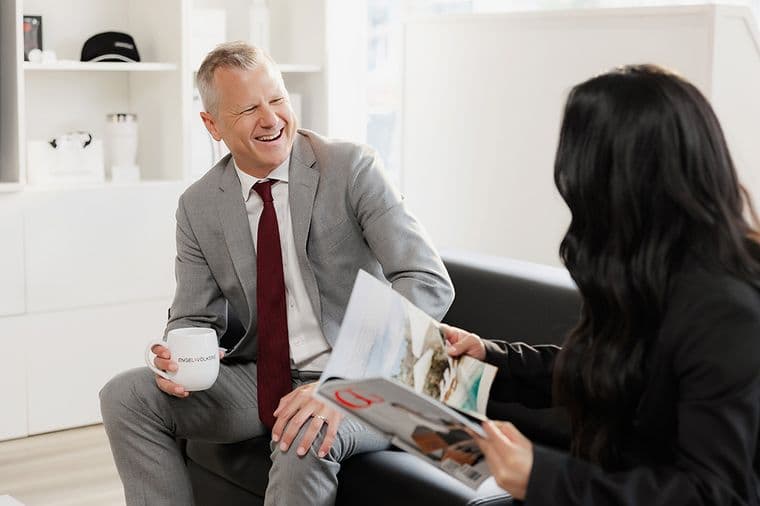 A man in a gray suit, holding a mug, smiles at a woman reading a magazine. They're seated in a modern, bright office space.