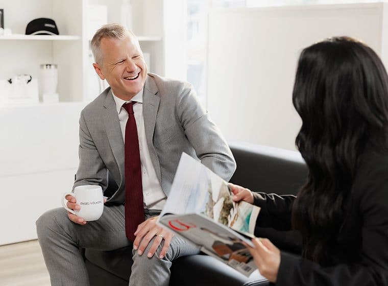 A man in a gray suit, holding a mug, smiles at a woman reading a magazine. They're seated in a modern, bright office space.
