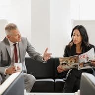A man and woman in business attire sit on a couch. The man gestures while holding a mug, and the woman reads a magazine.