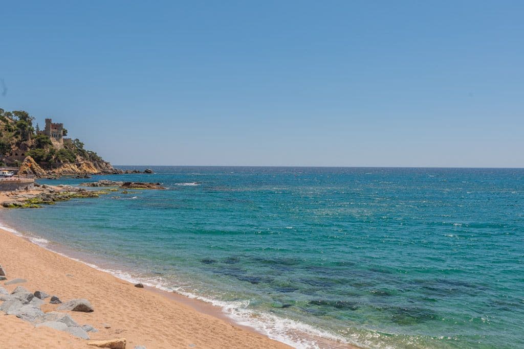 Platja de sorra es troba amb mar turquesa sota un cel blau clar. Un castell s'assenta al cim d'un penya-segat rocós a la distància.