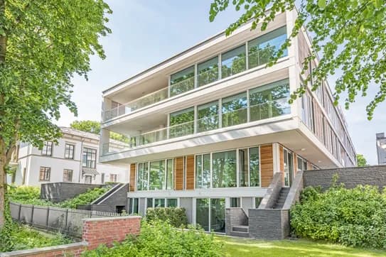 Modernes, dreistöckiges Wohngebäude mit Glasfassade, großzügigen Balkonen, weißer Fassade und Holzakzenten, eingebettet in Bäume und Grünanlagen.