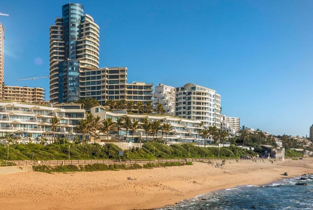 Panoramic view of Umhlanga Rocks beachfront in South Africa, featuring luxury high-rise apartment buildings and holiday flats overlooking the Indian Ocean. The scene showcases modern architecture with a distinctive cylindrical tower, palm trees lining the shore, and a sandy beach meeting the blue ocean waters. The coastline development is captured under clear blue skies on a sunny day.