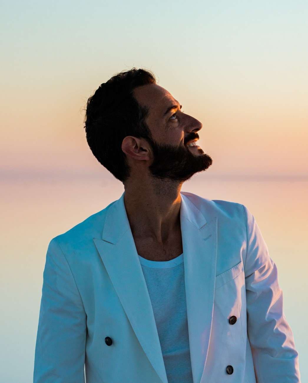Man in a white suit smiles, looking up at a sunset sky.