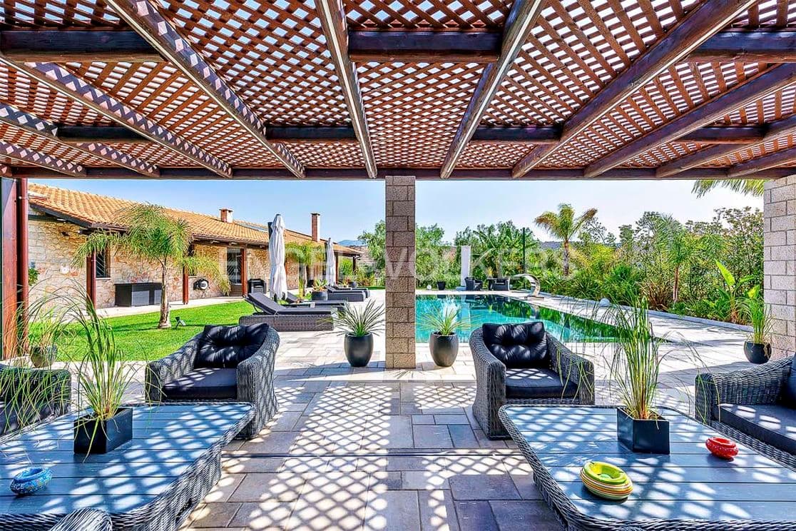 Outdoor patio with wicker furniture, a pool, and a wooden pergola. Lush greenery and a stone house are in the background.