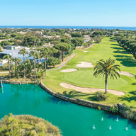 Luftaufnahme eines üppigen Golfplatzes mit Sandbunkern, Palmen und einem türkisfarbenen Wasserelement vor dem Hintergrund von Häusern und dem Meer.