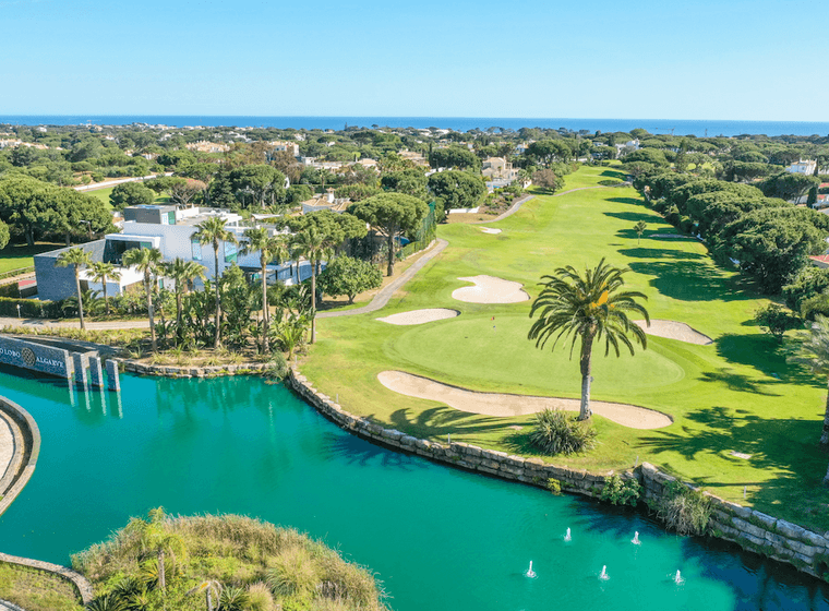 Luftaufnahme eines üppigen Golfplatzes mit Sandbunkern, Palmen und einem türkisfarbenen Wasserelement vor dem Hintergrund von Häusern und dem Meer.