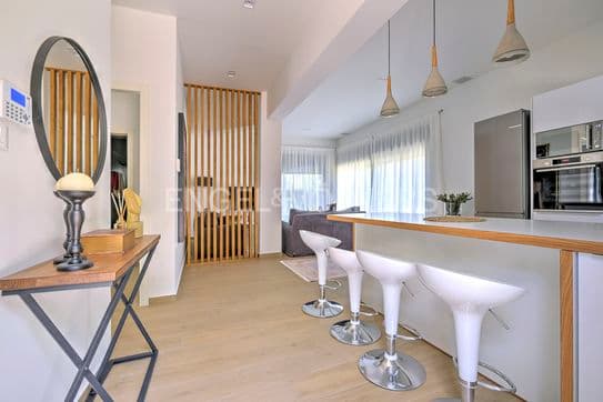 A bright, modern interior with a wood floor, white walls, and an open kitchen with white stools at the counter. A wooden screen separates the living area.