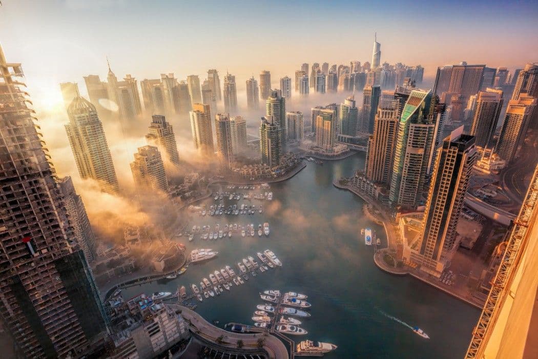 Aerial view of Dubai Marina at sunrise, showcasing high-rise residential towers, waterfront promenades, and yachts docked along the marina.