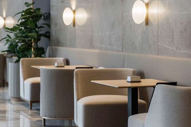 Modern cafe interior with beige chairs, wooden tables, green plant, and round wall lamps on marble walls, reflecting a stylish ambiance.