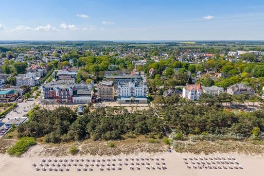 Luftaufnahme einer Küstenstadt mit Reihen von Strandliegen an einem Sandstrand, von Bäumen gesäumten Dünen und farbenfrohen Hotels und Häusern direkt am Strand