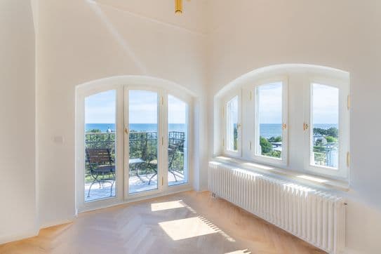 Ein heller, leerer Raum mit gewölbten Glastüren und Fenstern, die auf einen Balkon mit Stühlen und Meerblick führen; Sonnenlicht fällt auf den Fischgrätenparkettboden