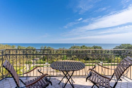 Balkon mit Holztisch und -stühlen sowie einem kunstvoll verzierten Eisengeländer, mit Blick auf die Baumkronen und das ruhige blaue Meer