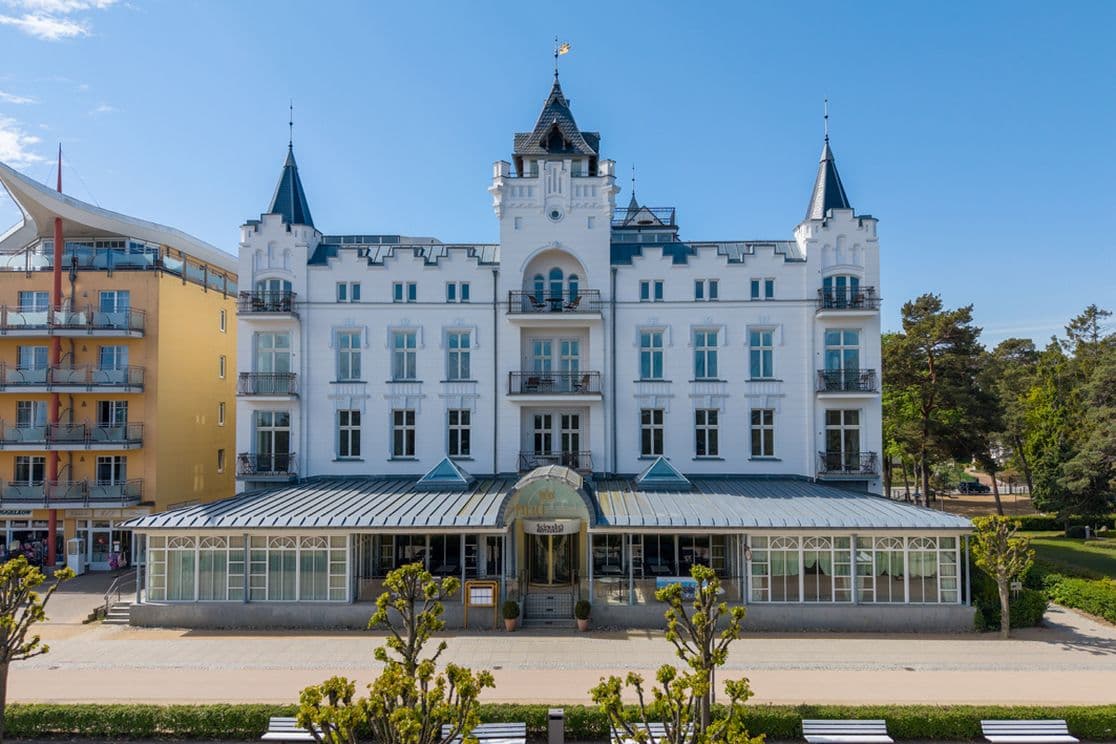 Ein weißes, historisches Hotel am Meer mit Türmchen und Balkonen, einem Erdgeschoss mit Glasfassade, gepflegten Bäumen und Bänken entlang der Promenade