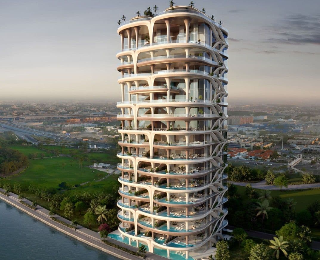 Futuristic high-rise building with unique curved architecture, surrounded by greenery and overlooking a body of water at sunset.