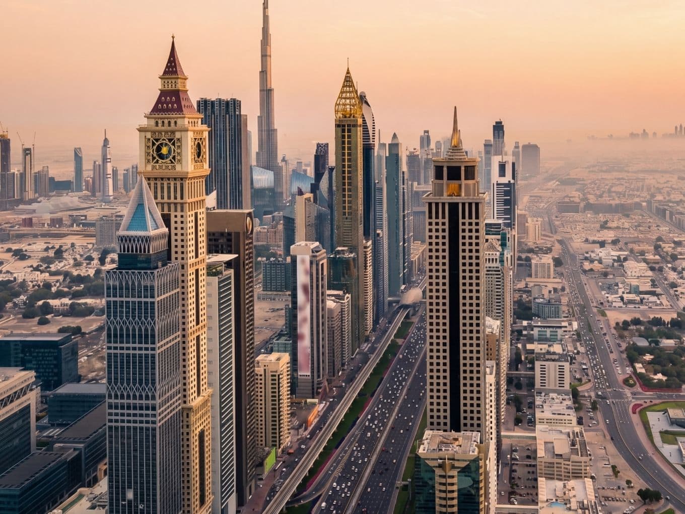 Aerial view of Dubai's skyline at sunset, featuring iconic skyscrapers and busy streets under a warm, hazy sky.