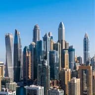 A dense cluster of glassy modern skyscrapers rises along a canal under a clear blue sky.