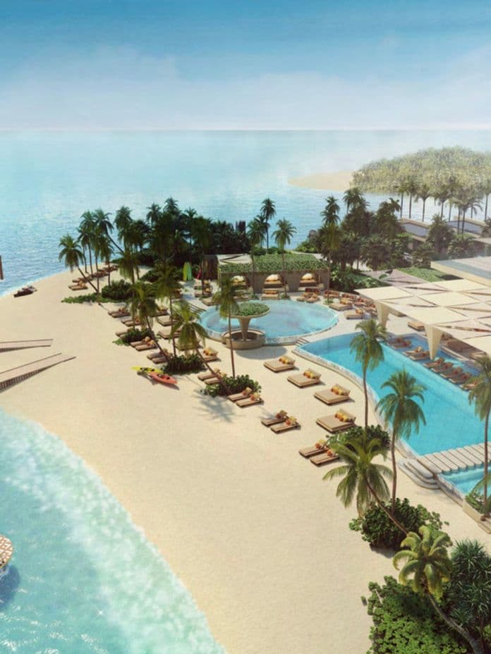 Aerial view of a tropical beach resort with palm trees, lounge chairs, pools, and clear blue ocean under a sunny sky.