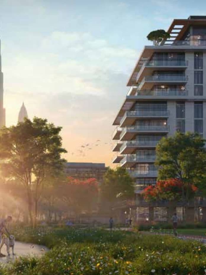 Modern apartment building with balconies, surrounded by greenery and a walkway. Tall skyscraper visible in the background at sunset.