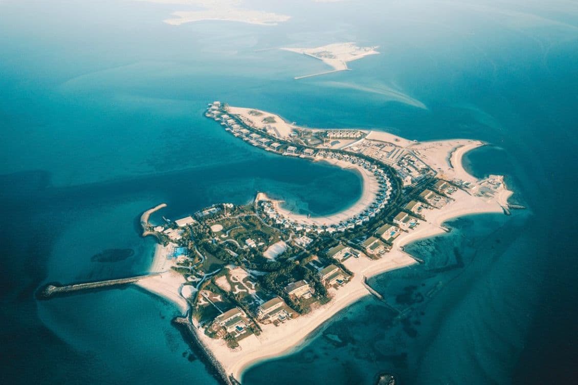 Aerial view of a luxurious island resort with villas surrounded by turquoise ocean waters and sandy beaches, under a clear blue sky.