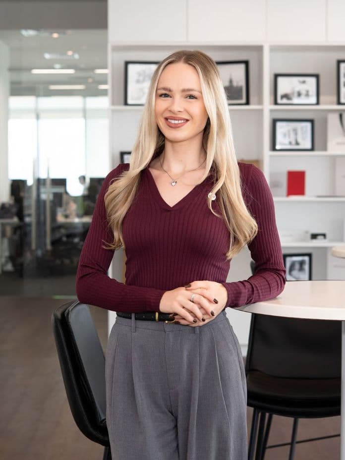 Smiling woman with long blonde hair in a burgundy top and gray pants, standing in a modern office with framed photos on shelves.